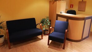 A reception area with a wooden desk, two chairs, a potted plant, and a sofa. - Hostal Hispanico II (Salamanca)