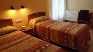 A hotel room with two single beds, a nightstand with a remote control, a wall socket, and a radiator.