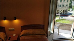 A room with a bed, a nightstand, a wall-mounted light, and a sliding glass door leading to an outdoor area with a balcony and a building in the background. - Hostal Hispanico II (Salamanca)