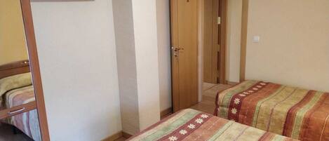 Una habitación pequeña y funcional con dos camas individuales, un armario de madera y una puerta que da a otra habitación.