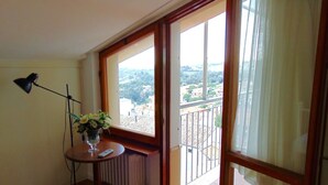 A room with a radiator, a table with a vase of flowers, a standing lamp, and a large window with a view of buildings and greenery. - CASA DEL SOLE - Numana, wonderful sea view penthouse (Numana)