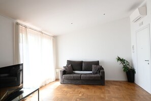 A modern living room with a grey sofa, a TV on a glass table, and a potted plant.