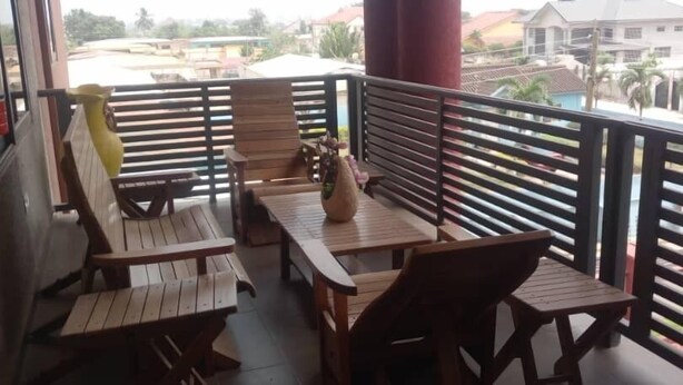 A covered outdoor seating area with wooden tables and chairs, a vase, and a view of buildings and a body of water in the background. - Alpha's Court (Kumasi)