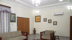 A living room with a sofa, armchair, ottoman, and a small table. There is a painting on the wall and an air conditioning unit mounted on the wall. - Alpha's Court (Kumasi)