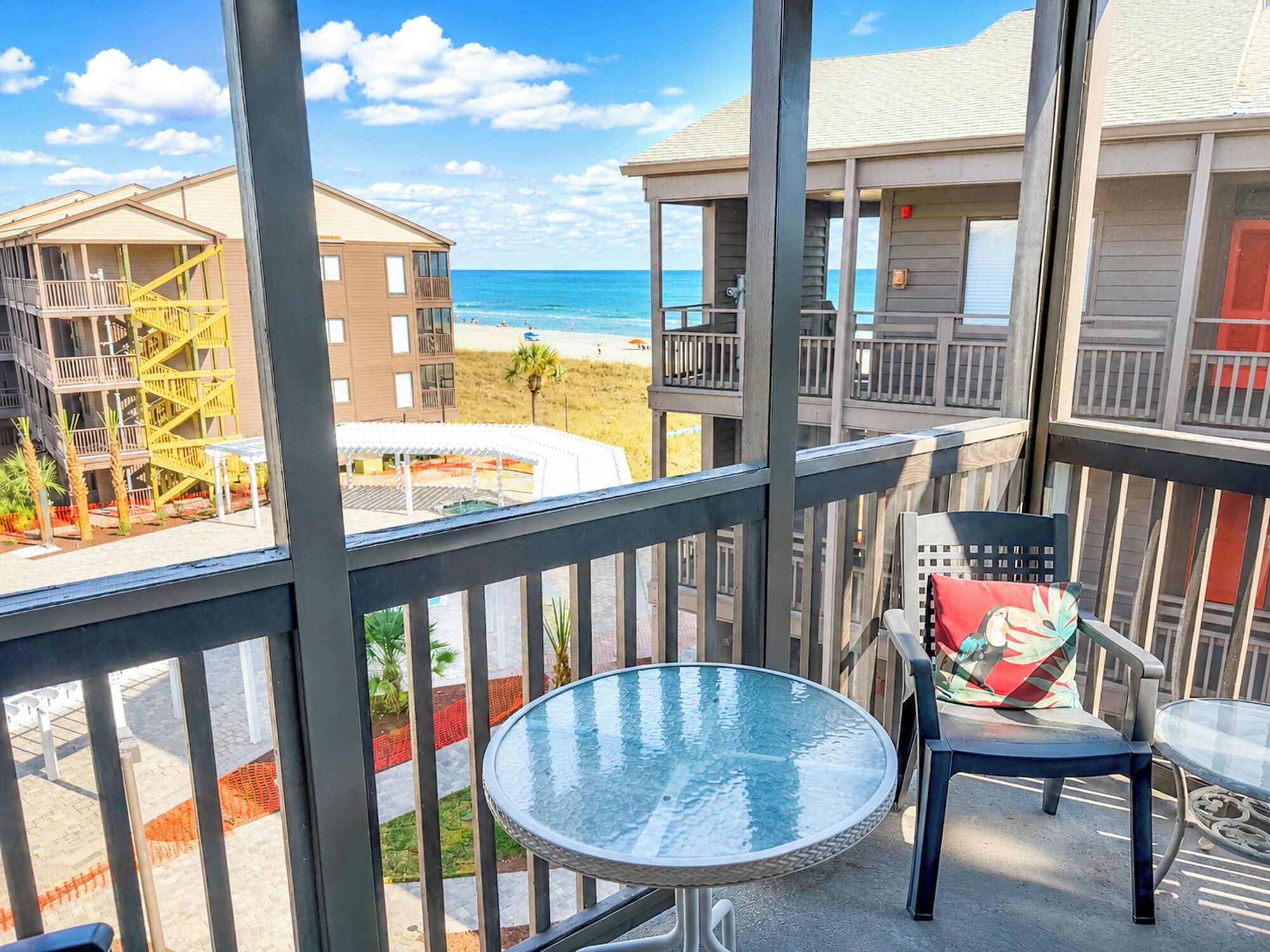 A balcony with a glass table and chairs, overlooking a beach and buildings.