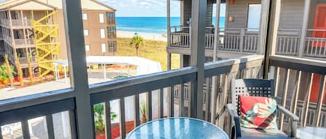 A balcony with a glass table and chairs, overlooking a beach and buildings.