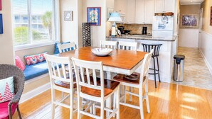 A compact kitchen with white cabinets, a dining table with chairs, a window with blinds, and a small trash can. - Experience Tilghman Beach and Racquet Club Unit 309! 3 Bedroom Ocean View Condo. Pools, Tennis, Close to Main Street! (North Myrtle Beach)
