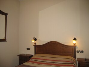 A neatly made bed with a striped blanket, two bedside lamps, a wooden headboard, a nightstand with a glass, and a wall-mounted mirror. - Hostal Granada (Salamanca)