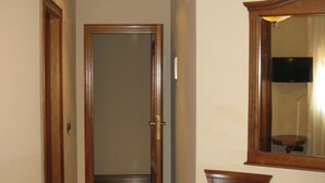 Un corridoio stretto con una porta in legno in fondo, una sedia con cuscino decorato e uno specchio appeso al muro.