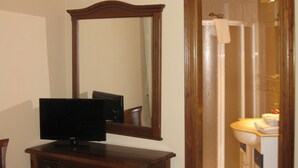 Una stanza con una scrivania in legno, una televisione, una sedia, uno specchio e un bagno visibile attraverso una porta aperta.