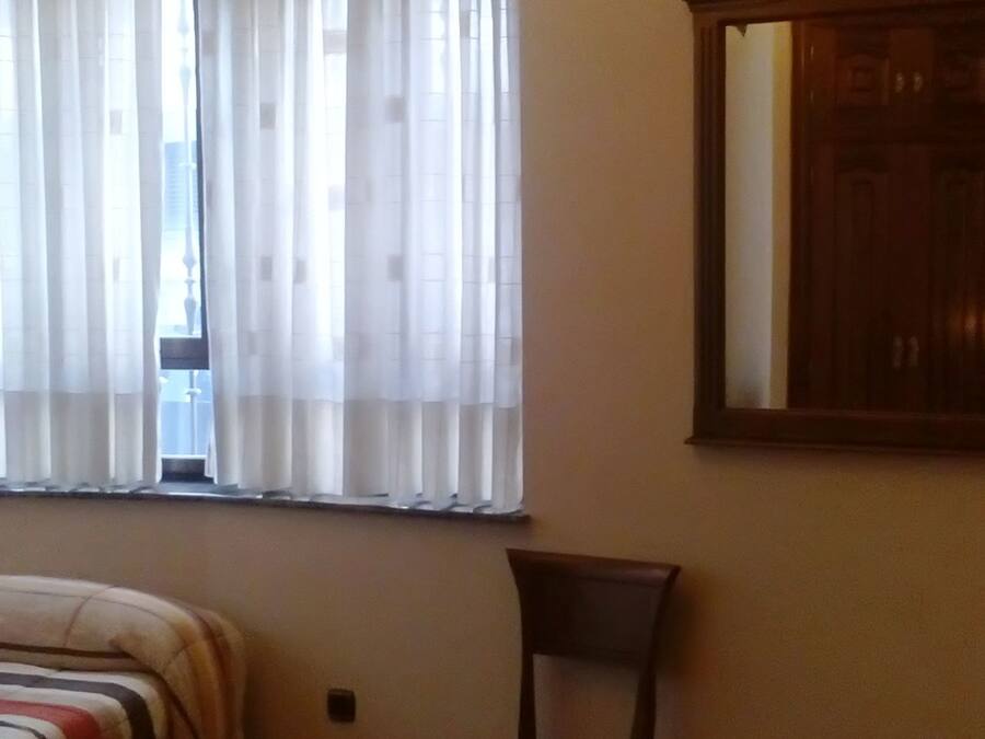 A bedroom with a bed, a chair, a wooden cabinet, and a window with curtains.