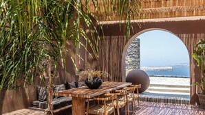 An outdoor seating area with a wooden table and chairs, a stone pillar, and a view of the sea. - Arota Exclusive Villas (Santorini)