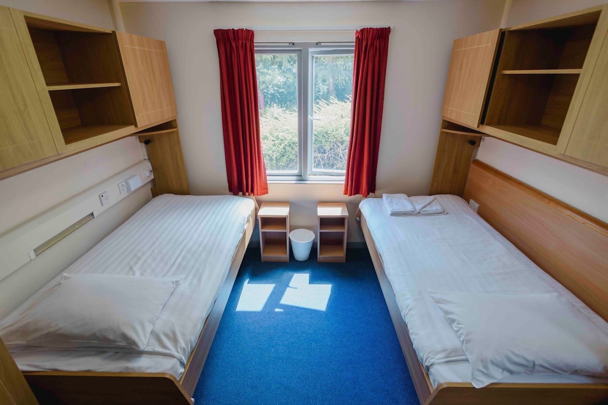 Une chambre avec deux lits, une fenêtre avec des rideaux, une petite table et une poubelle.