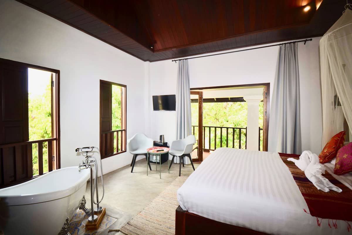 A hotel room with a large bed, a bathtub, a small table with chairs, and a balcony with curtains.