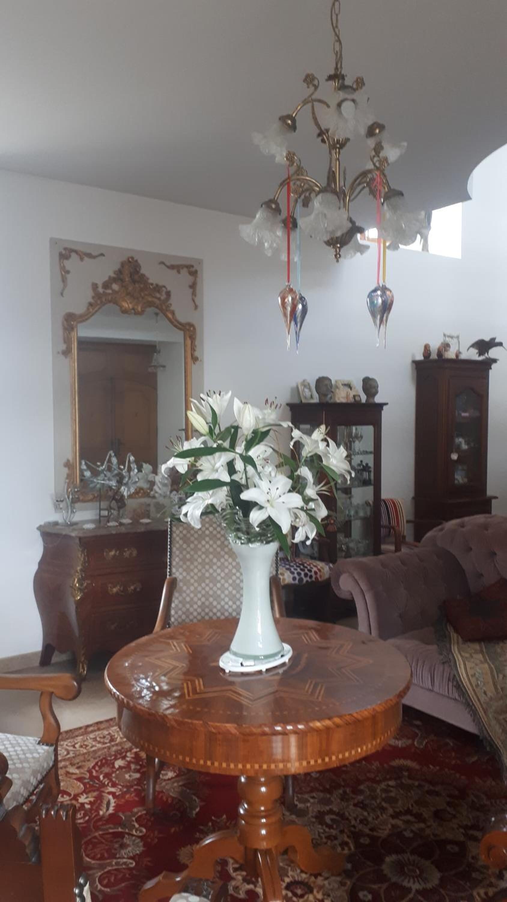 Un salon avec une table ronde en bois, un vase blanc orné de fleurs, un grand miroir et un lustre.