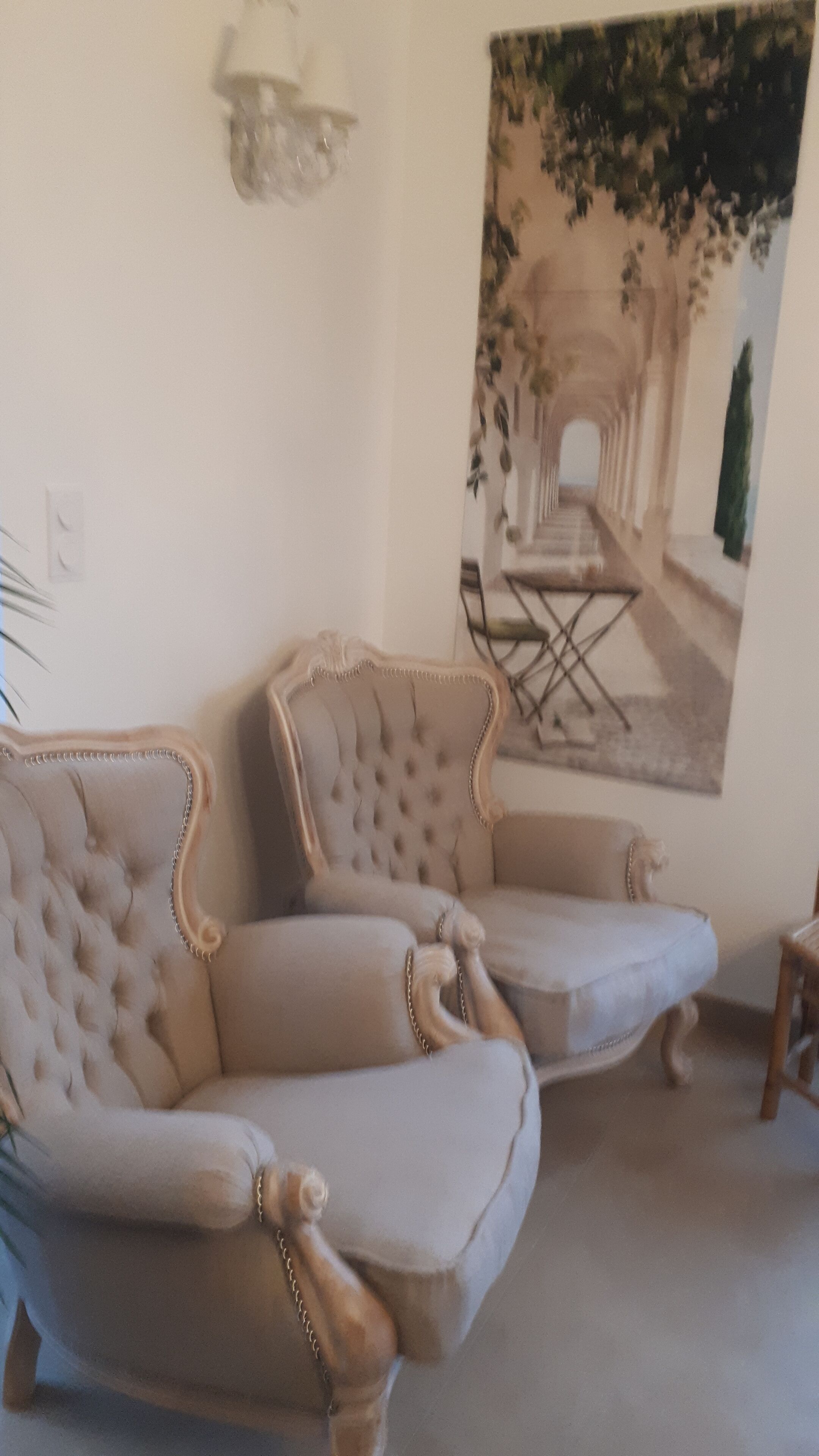 A room with a beige upholstered armchair and a matching ottoman. There is a painting of an outdoor scene on the wall.