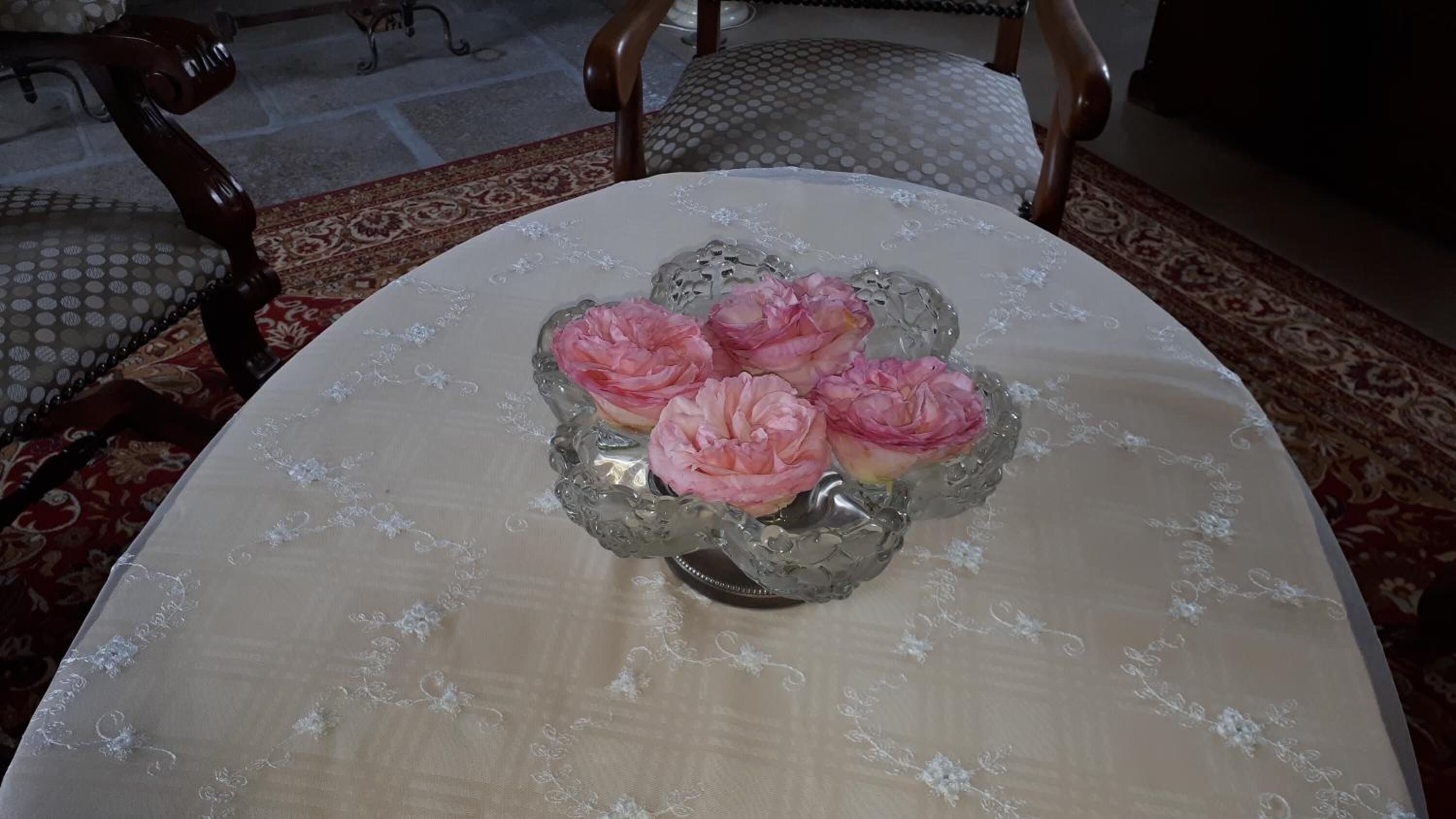 A round table with a floral tablecloth and a glass vase containing pink flowers.
