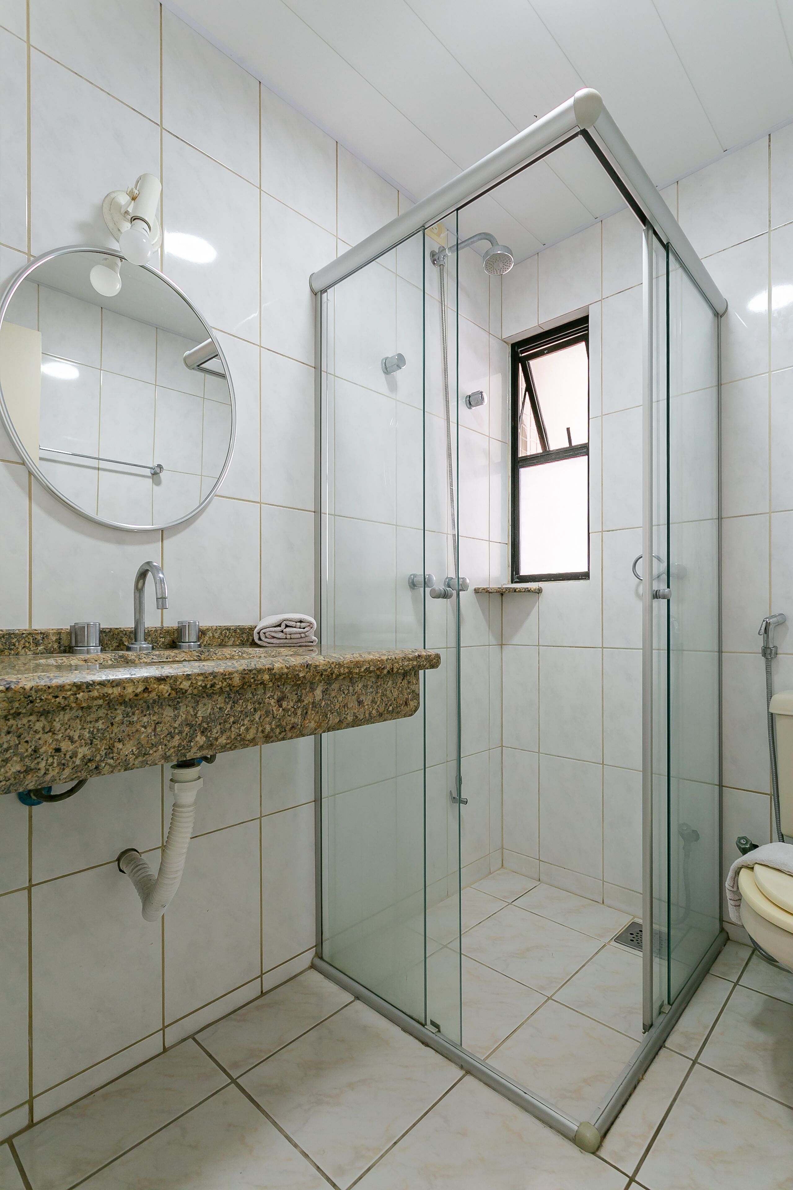 Un baño con ducha envuelta en vidrio, lavamanos de encimera de granito, espejo redondo y un inodoro.
