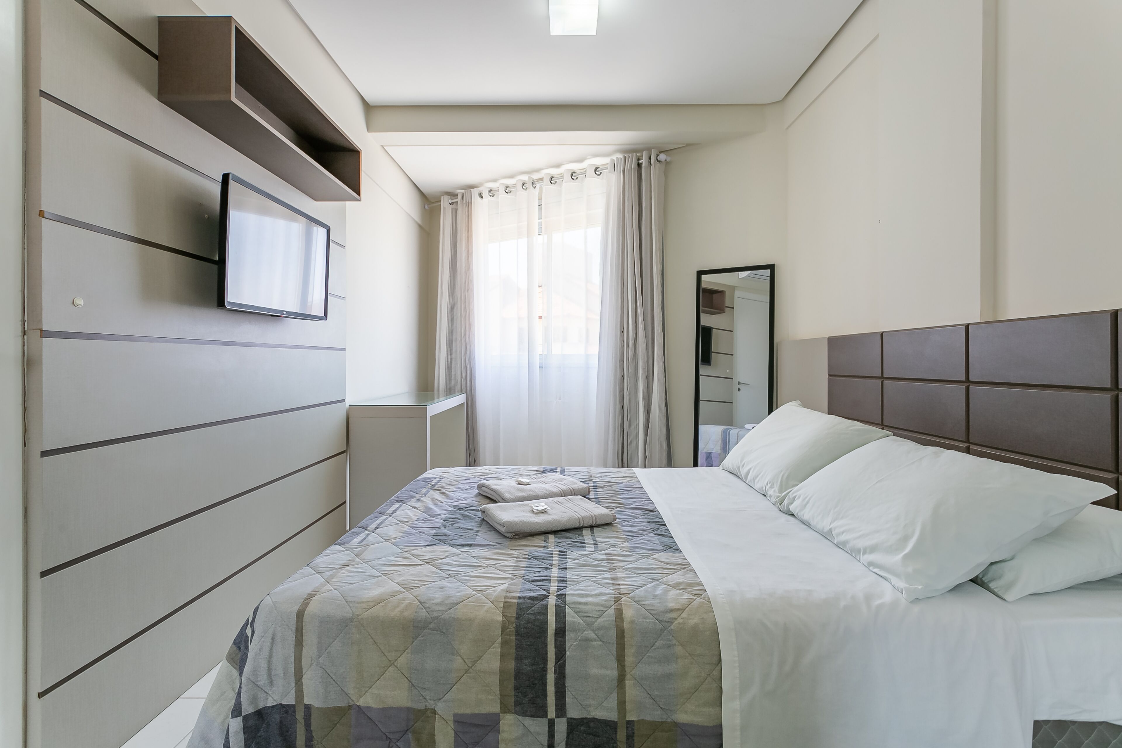 A modern hotel room with a bed, a flat-screen TV mounted on the wall, a large mirror, and a window with sheer curtains.