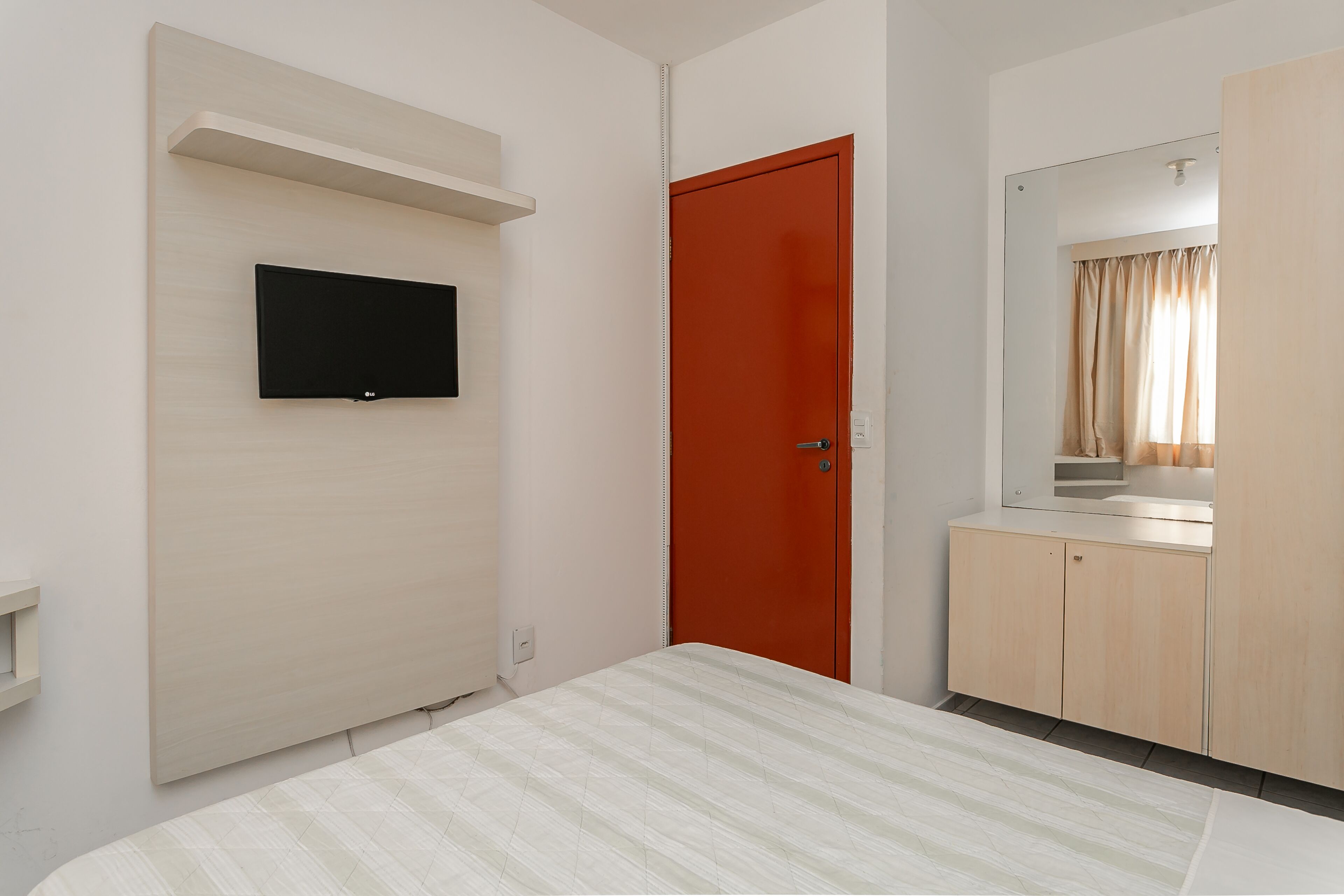 Un dormitori amb llit, televisió penjada a la paret, porta vermella i armari de fusta.