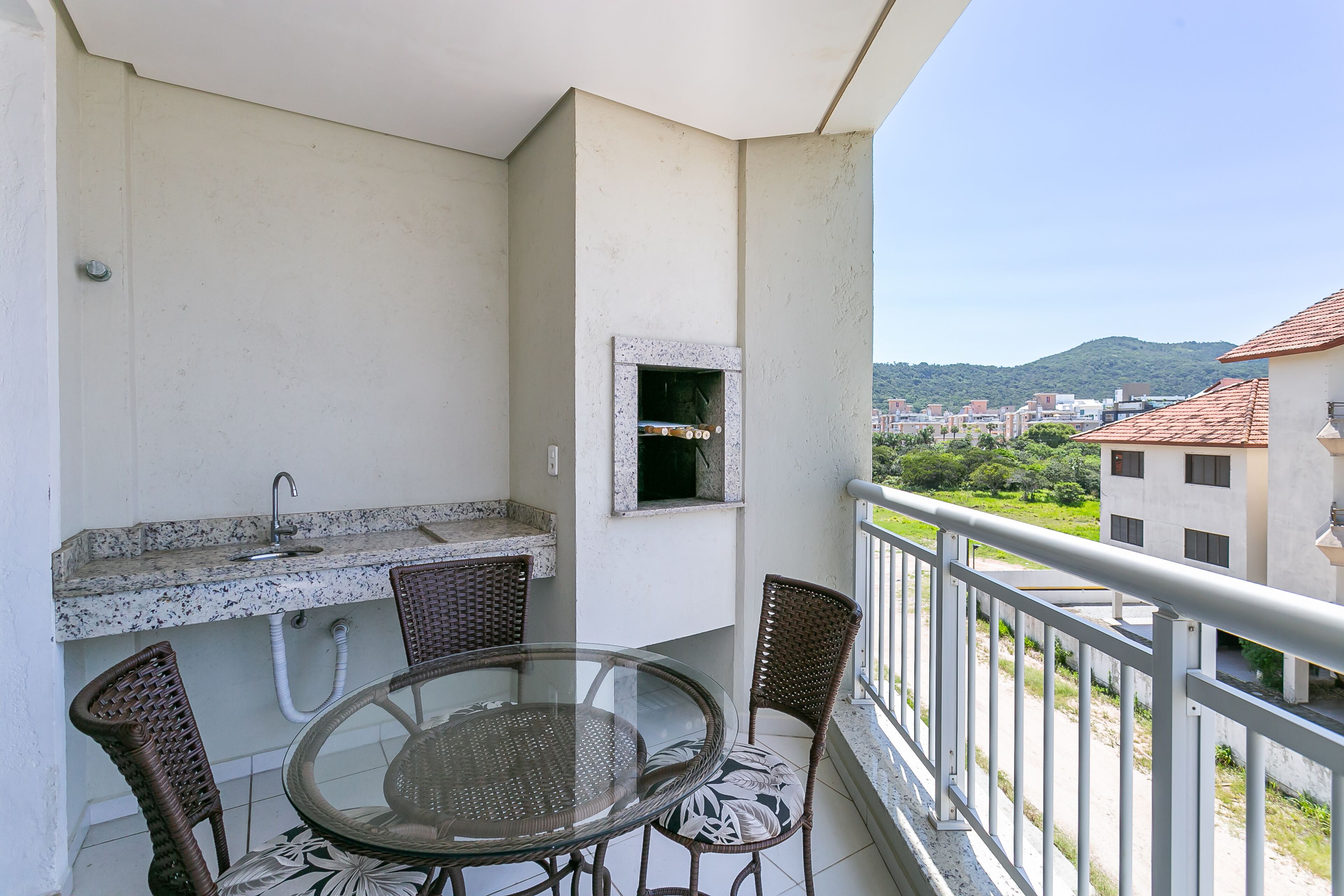 A balcony with a glass table and wicker chairs, a small kitchen area, and a view of a cityscape and hills in the background.