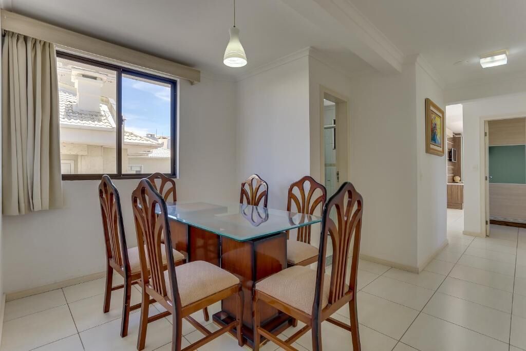 A dining area with a glass table and wooden chairs, a window with curtains, and a hallway leading to another room.
