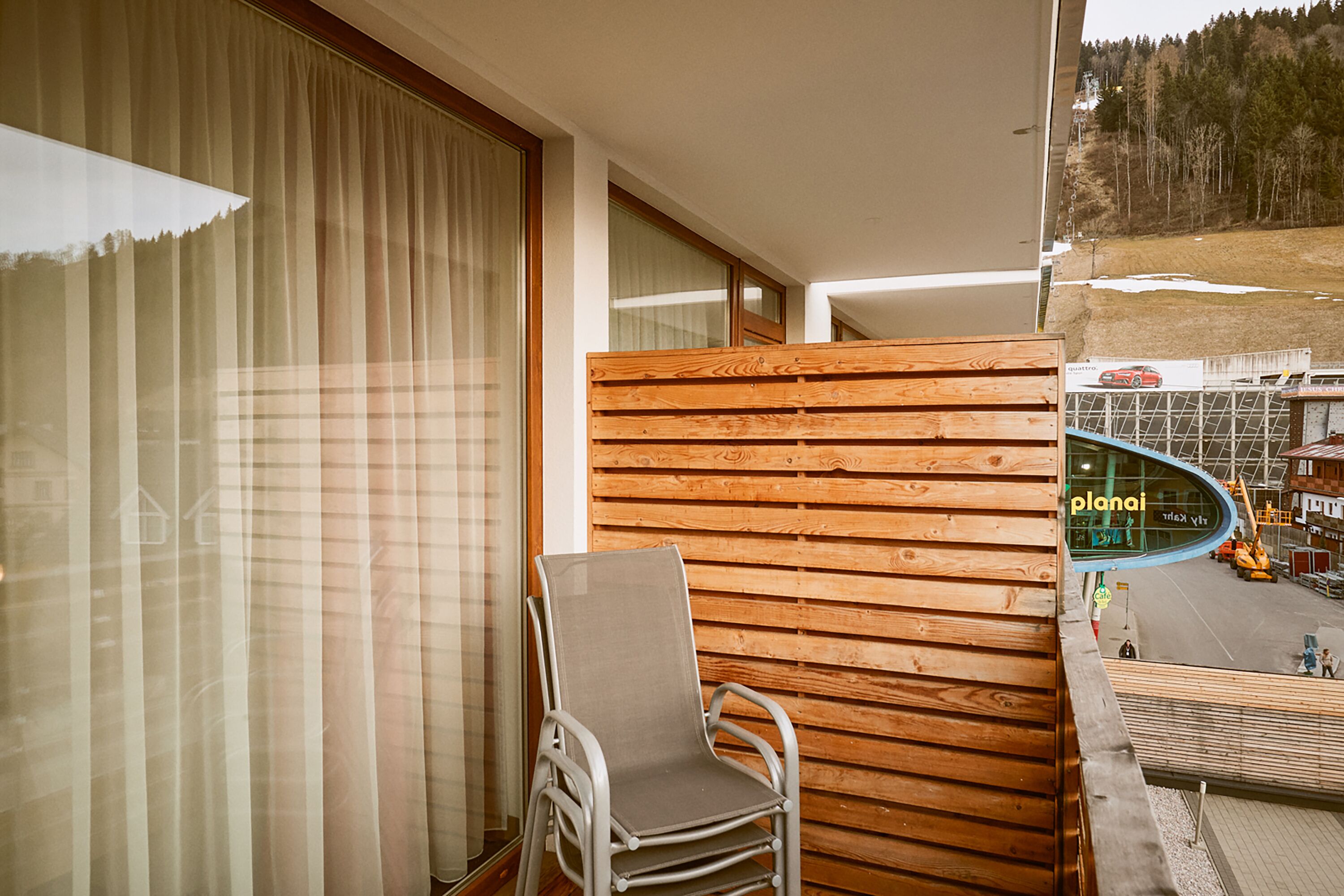 A balcony with a wooden partition, a chair, and a view of a road with vehicles and a building labeled "planai".
