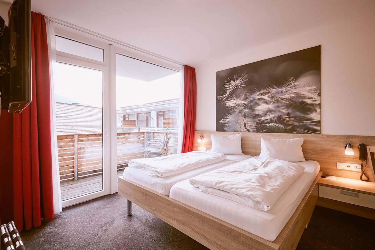 A hotel room with a large bed, a sliding glass door to the outside, a painting on the wall, and a bedside table with a lamp.