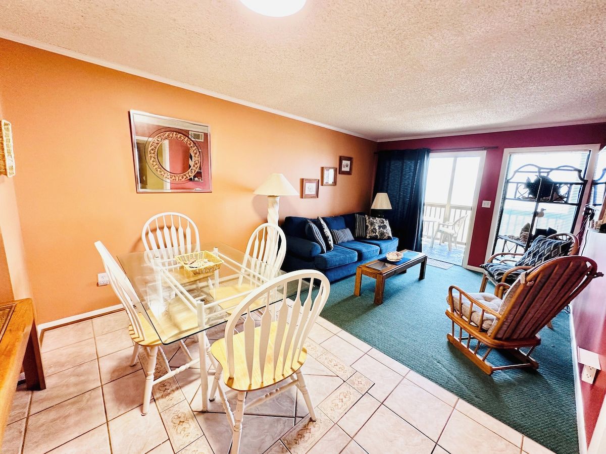 A living room with a glass dining table, white chairs, a blue sofa, and a rocking chair.
