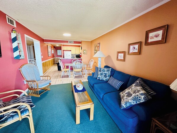 A living room with a blue sofa, wooden chairs, a small wooden table, and a kitchen in the background. - Experience Tilghman Beach and Racquet Club Unit 122! Oceanfront, Pools, Tennis, Close to Main Street! (North Myrtle Beach)