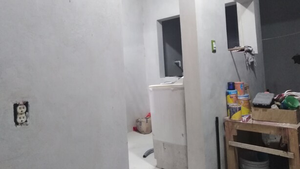 Ein Zimmer mit weißer Wand, einem Fenster und einer Tür. An der Holzwand befindet sich ein Regal mit verschiedenen Gegenständen.