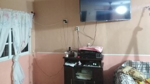 Ein Zimmer mit an der Wand montiertem Fernseher, einem Fenster mit Vorhängen, einem Bett mit gemustertem Bettbezug und einem kleinen Tisch mit verschiedenen Gegenständen.