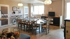 Zona pranzo con un tavolo grande, sedie e lampade a sospensione. Si intravede una bicicletta sullo sfondo e una TV su un mobile.