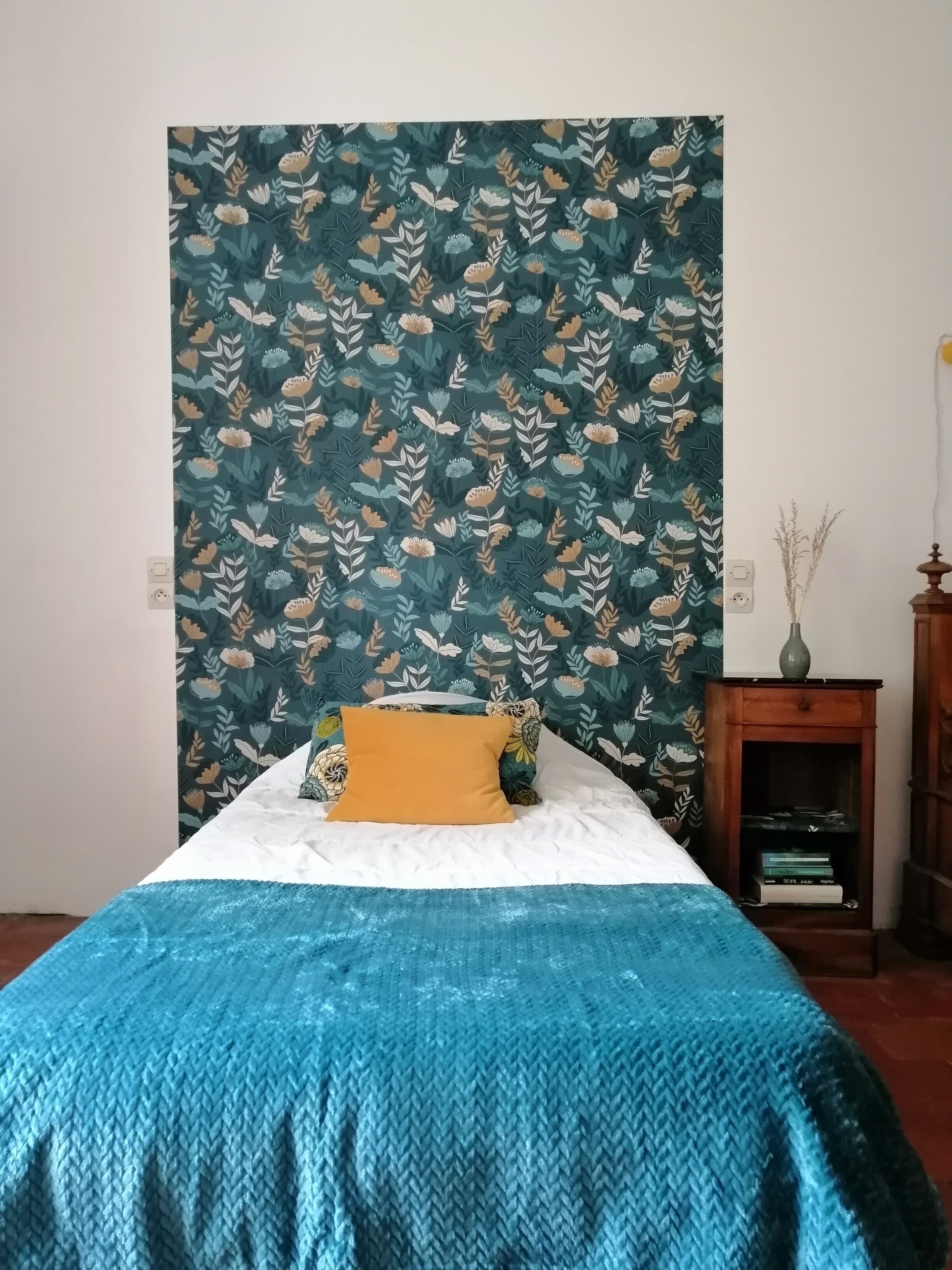 Un dormitori amb un llit, una manta blava amb textura, un tapís floral i una petita taula auxiliar de fusta amb un ram.