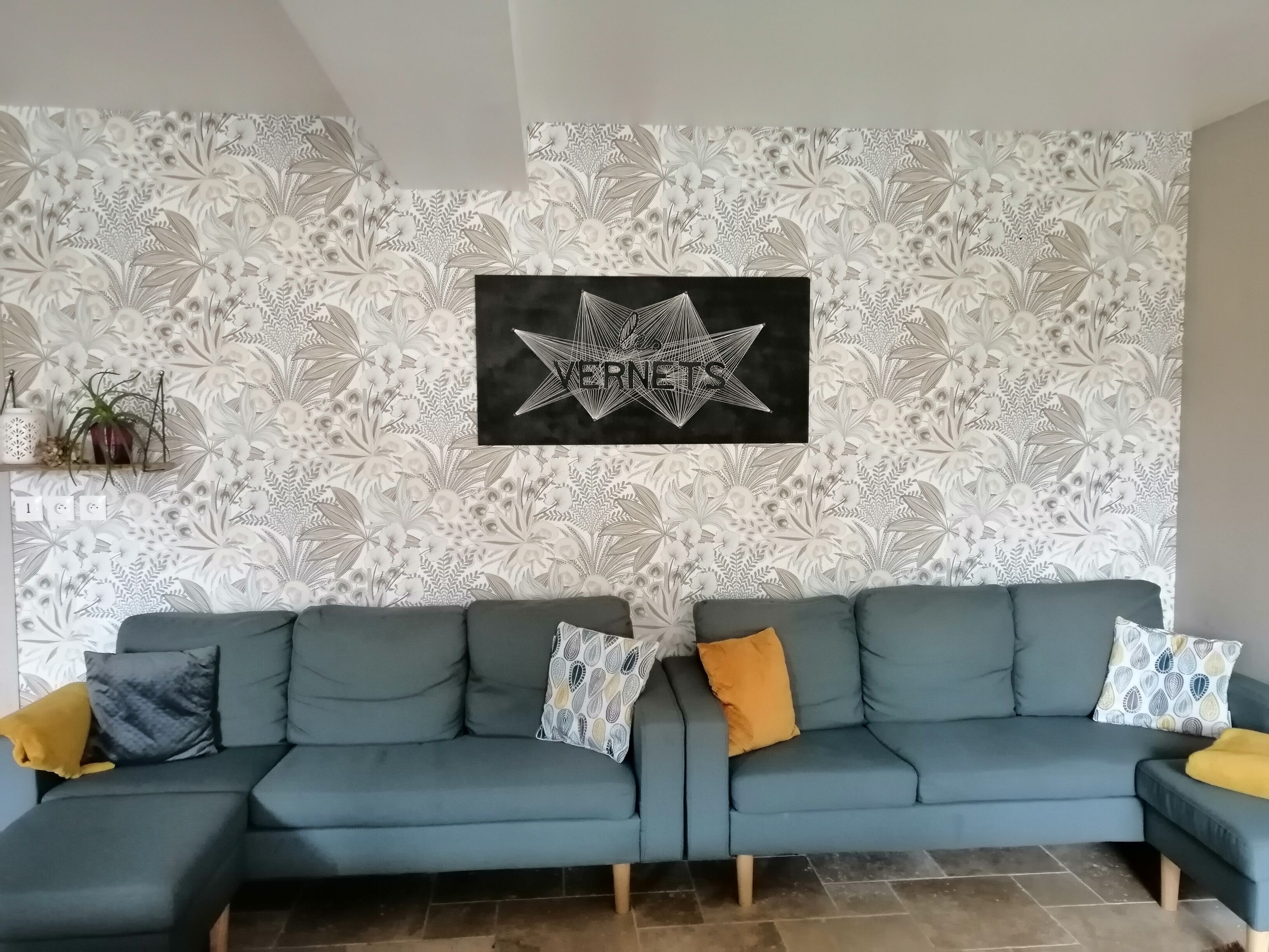 A living room with a sectional sofa, patterned wallpaper, and a framed artwork on the wall.
