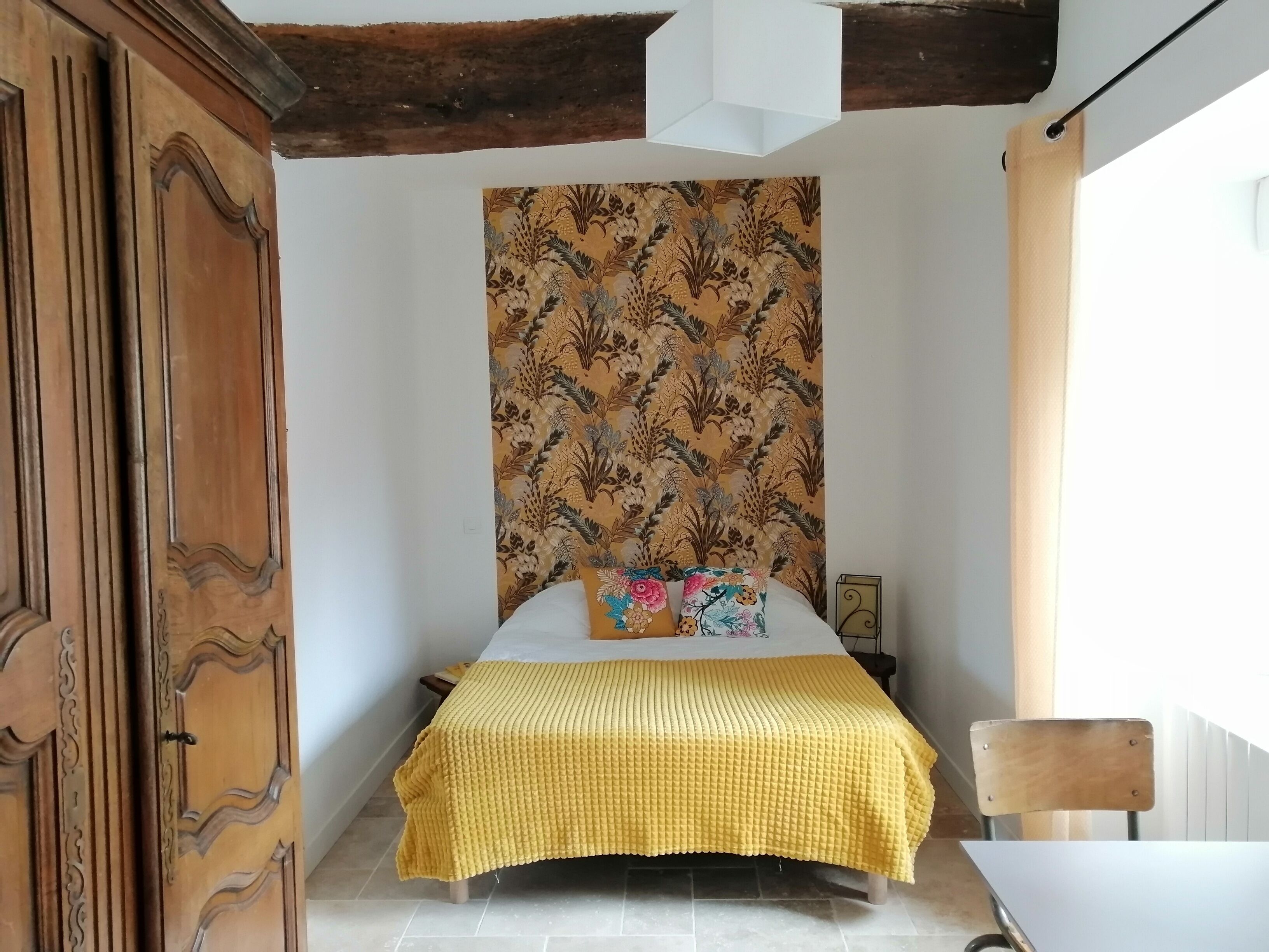 A bedroom with a bed, a wooden wardrobe, a patterned tapestry, and a small table with a chair.