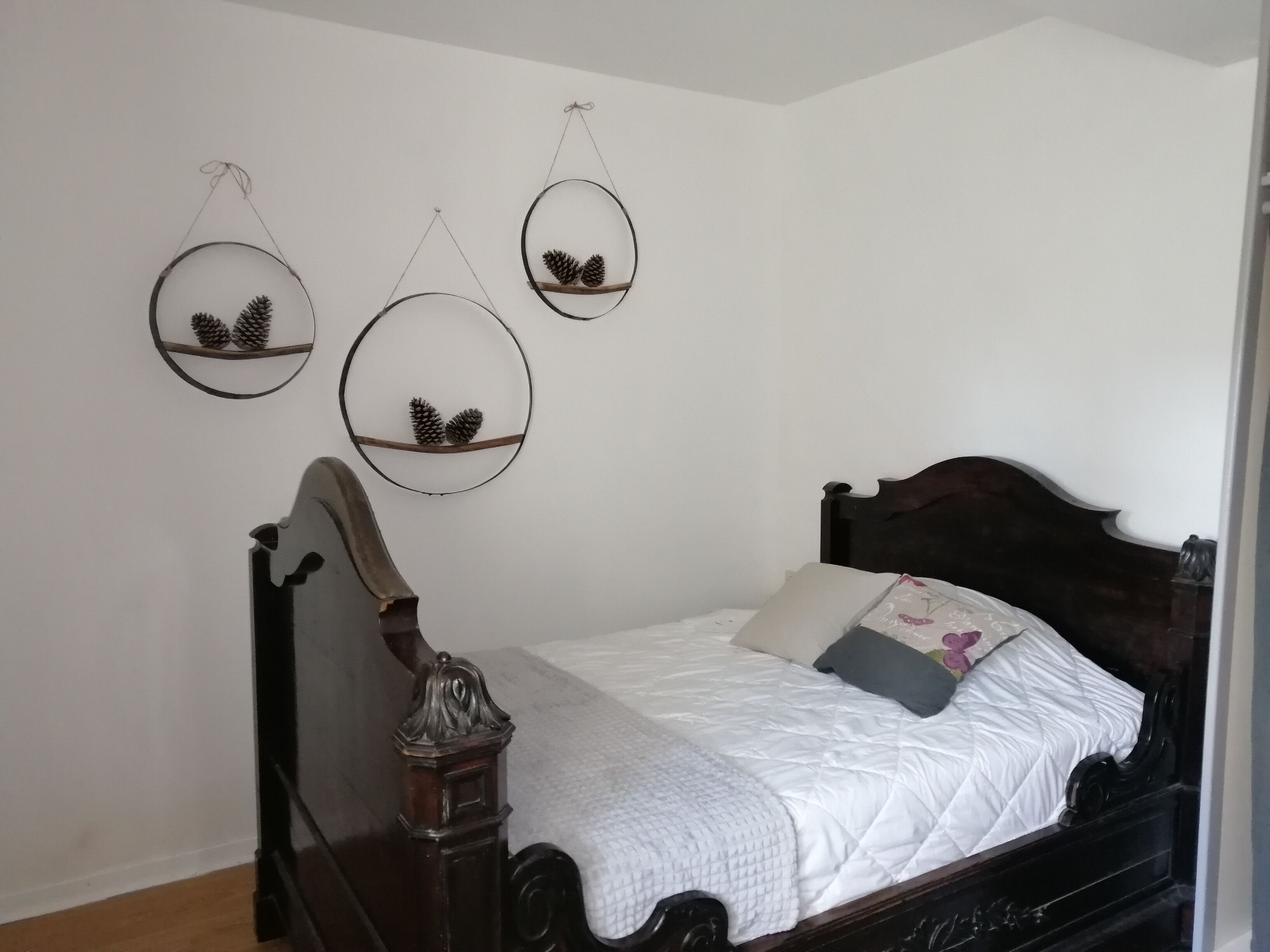 Un dormitorio con estructura de cama de madera oscura, ropa de cama blanca y tres estantes circulares fijos a la pared con piñas.