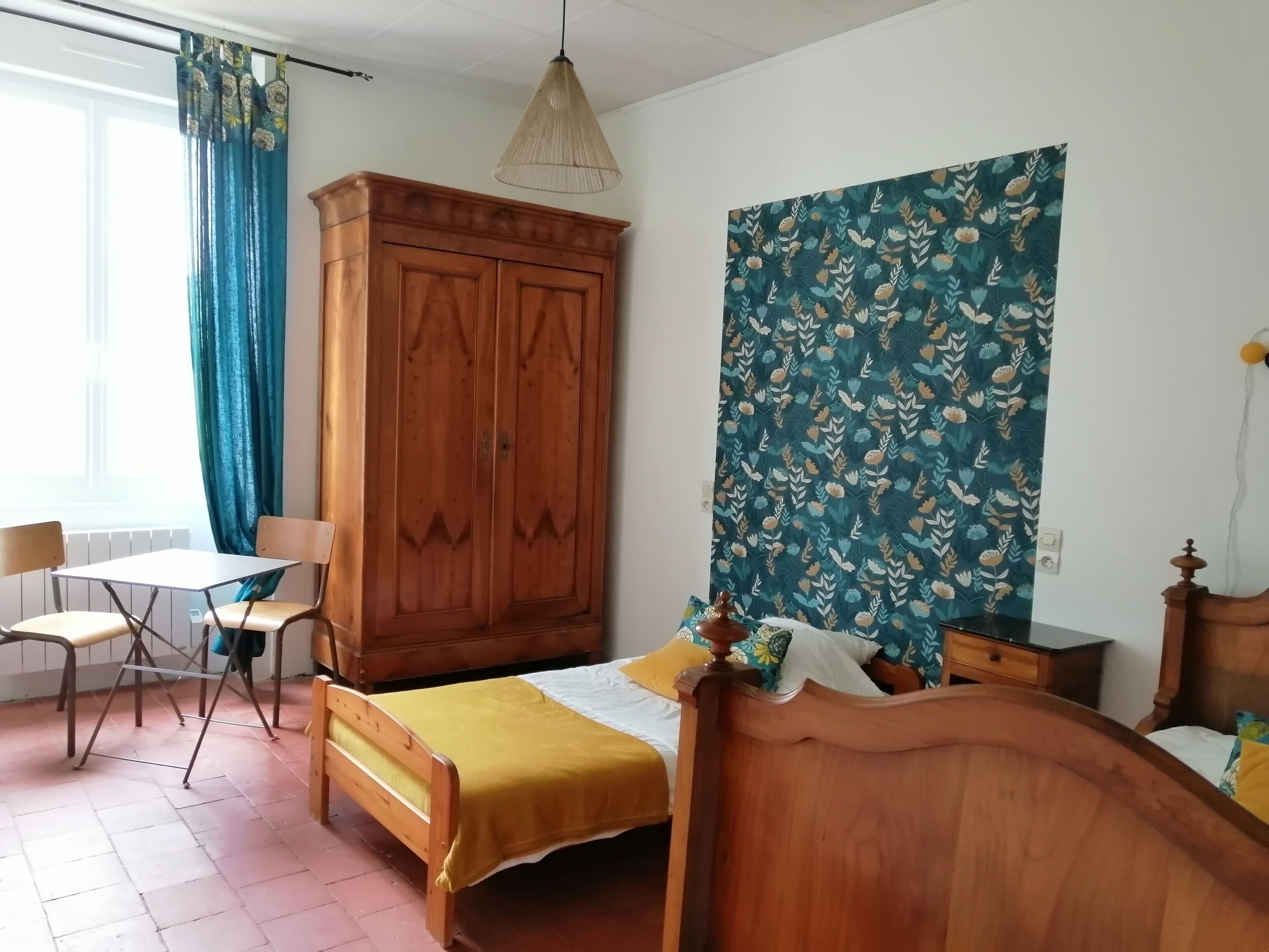 Une chambre à coucher avec un lit en bois, une armoire en bois, une petite table, une chaise et une grande tapisserie à motifs floraux accrochée au mur.