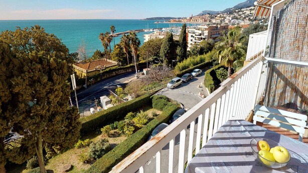 A balcony with a view of a coastal town, a road with parked cars, and a clear sky. - Top-floor 2-room with air con & private parking in Menton (Menton)