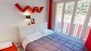 A bedroom with a bed, a window with a view of greenery, a red wall decoration, and a white headboard. - Top-floor 2-room with air con & private parking in Menton (Menton)