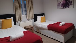 A hotel room with two beds, a window with curtains, a radiator, a nightstand, and a painting on the wall. - Huku Kwetu - Fieldfare Green (Luton)