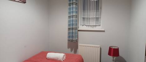Una camera da letto piccola e ordinata con un letto singolo, una finestra con tende leggere, un termosifone, un comodino con lampada e un quadro appeso al muro.