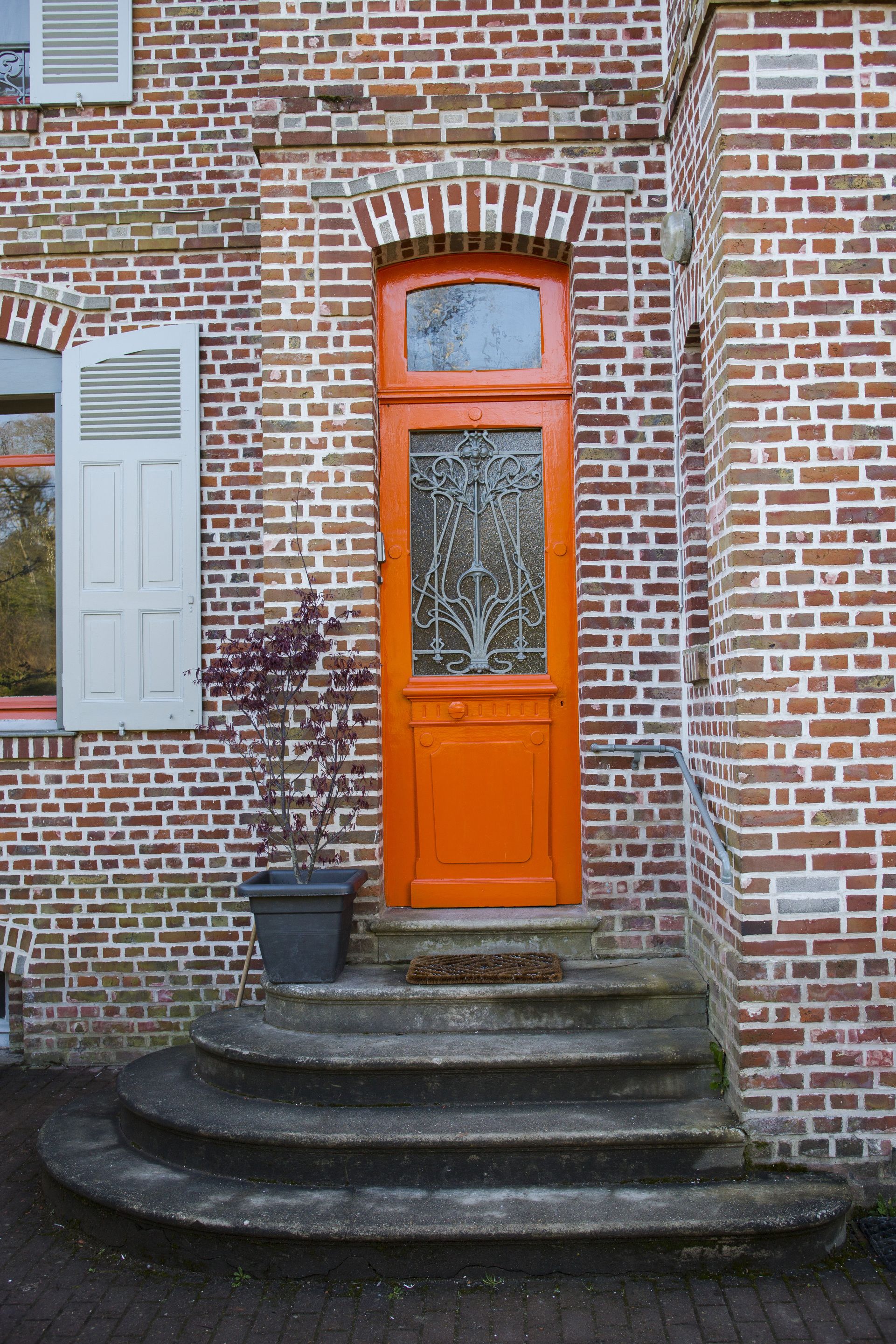 Een bakstenen gebouw met een rode deur, witte luiken en een bloempot.