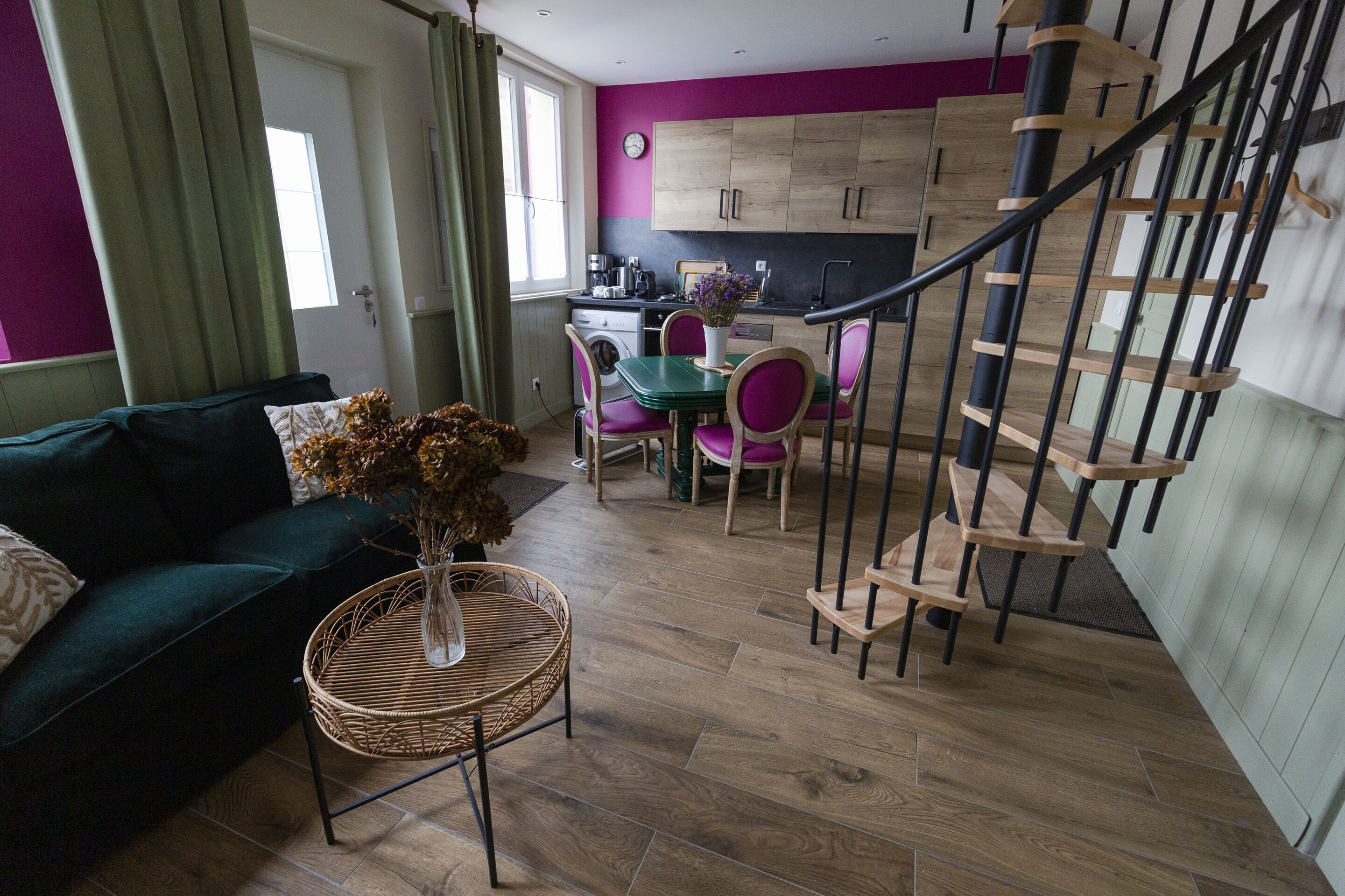 Un salon moderne avec un canapé vert, une table en osier ornée d’un vase et un coin repas avec une table verte et des chaises violettes. On aperçoit à l’arrière-plan une cuisine avec des meubles en bois et un escalier noir.