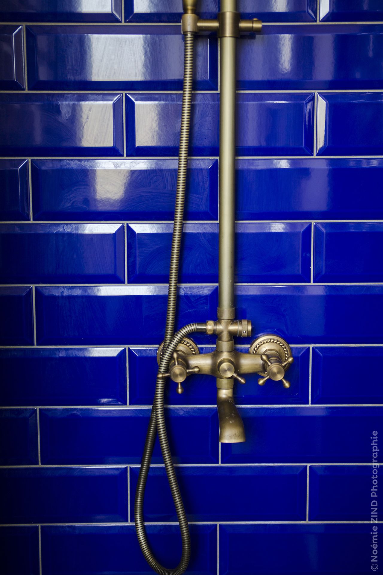 A shower with a hose and two valves against a blue tiled wall.