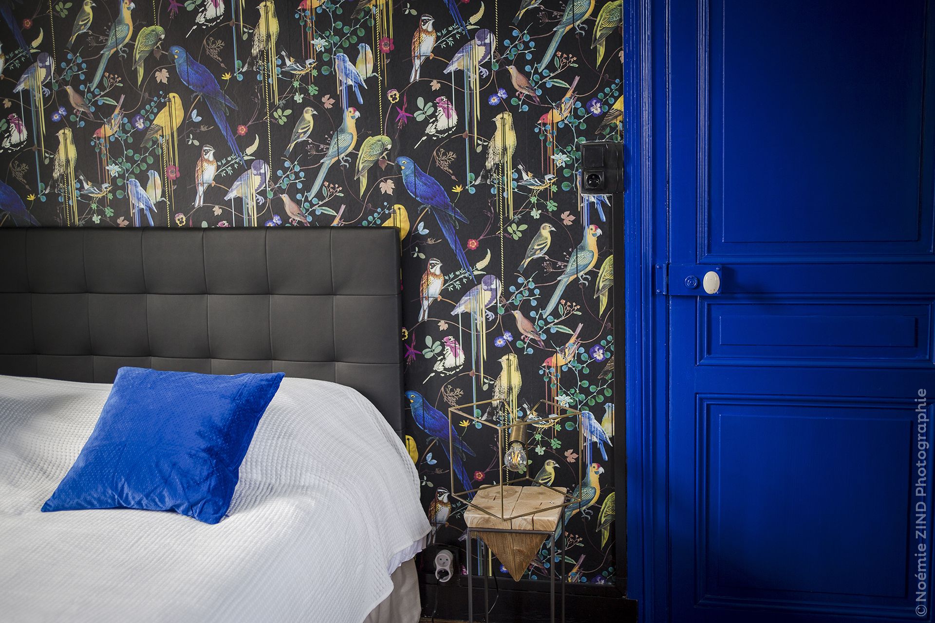 Un dormitorio con cabecera capitonada, cama con ropa de cama blanca y almohada azul, pared con mural de aves y flores, y una puerta azul.