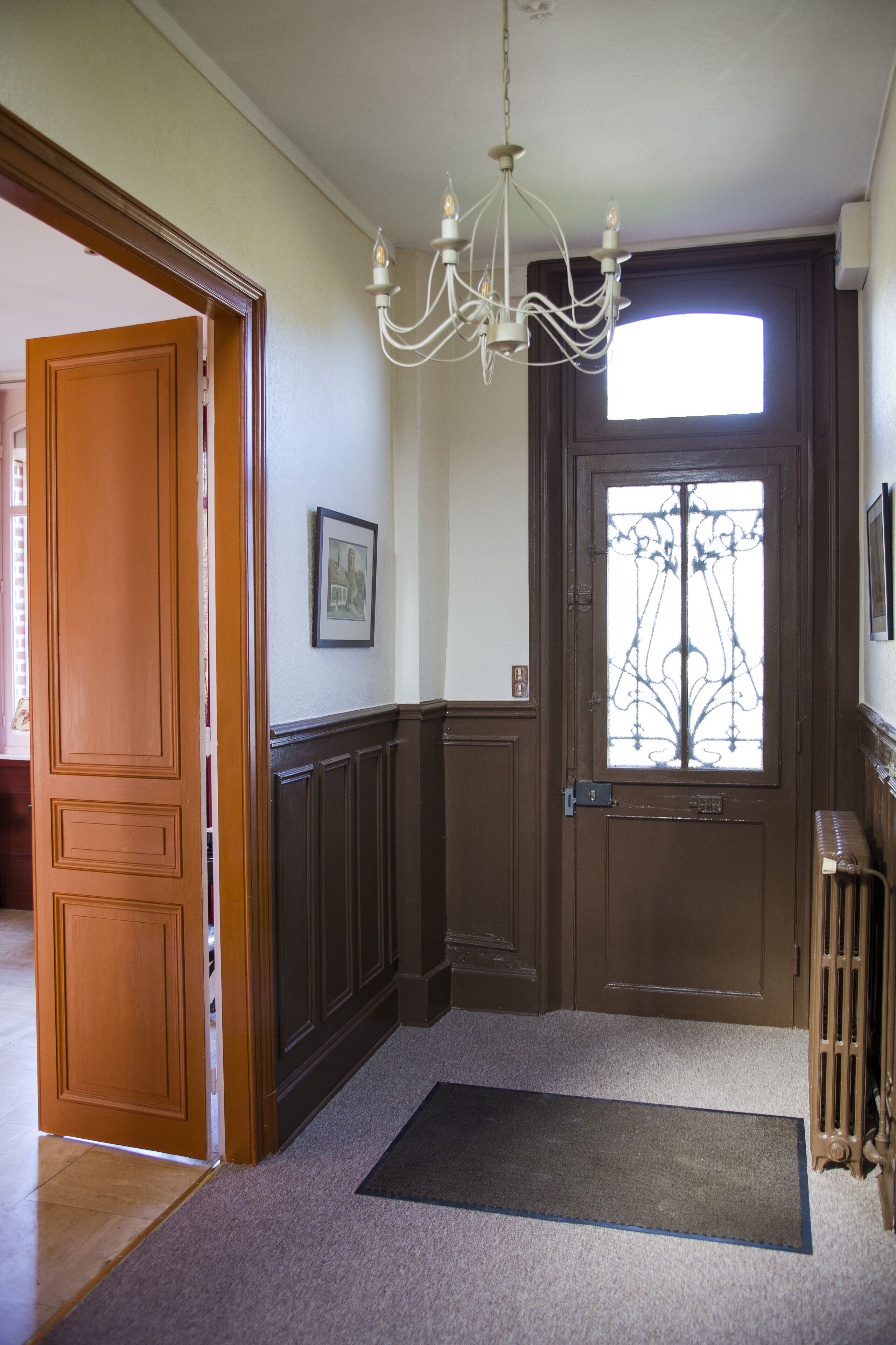 A hallway with a wooden door, a black doormat, a chandelier, and a radiator.