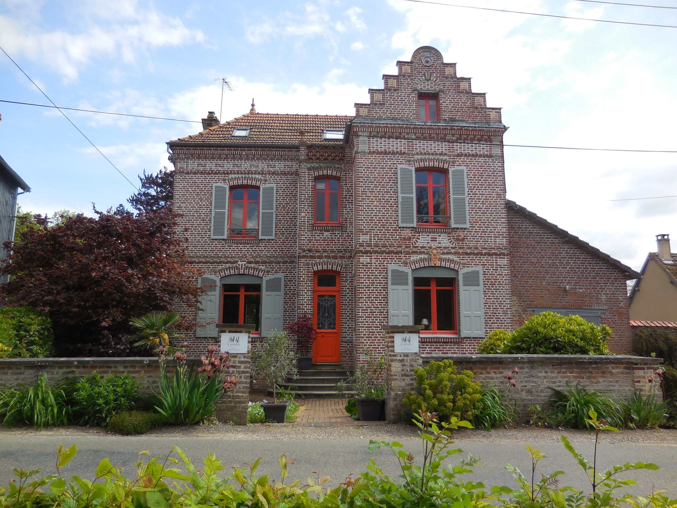 A two-story brick building with a red door and shutters, surrounded by greenery and a paved area.