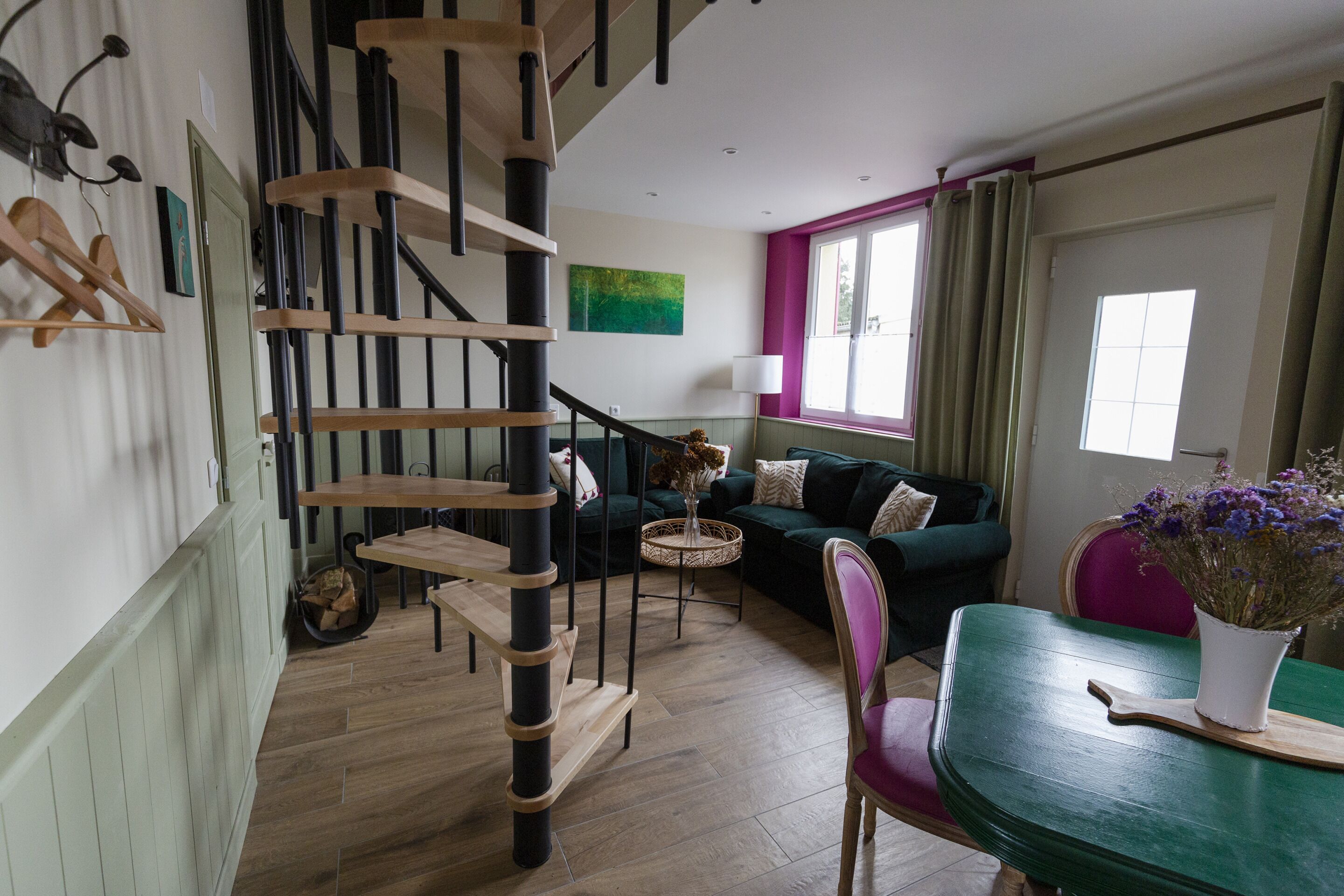 Un escalier avec des marches en bois et des rampes noires, un canapé vert, une petite table ronde avec un vase, et une table à manger ornée d’un vase de fleurs.
