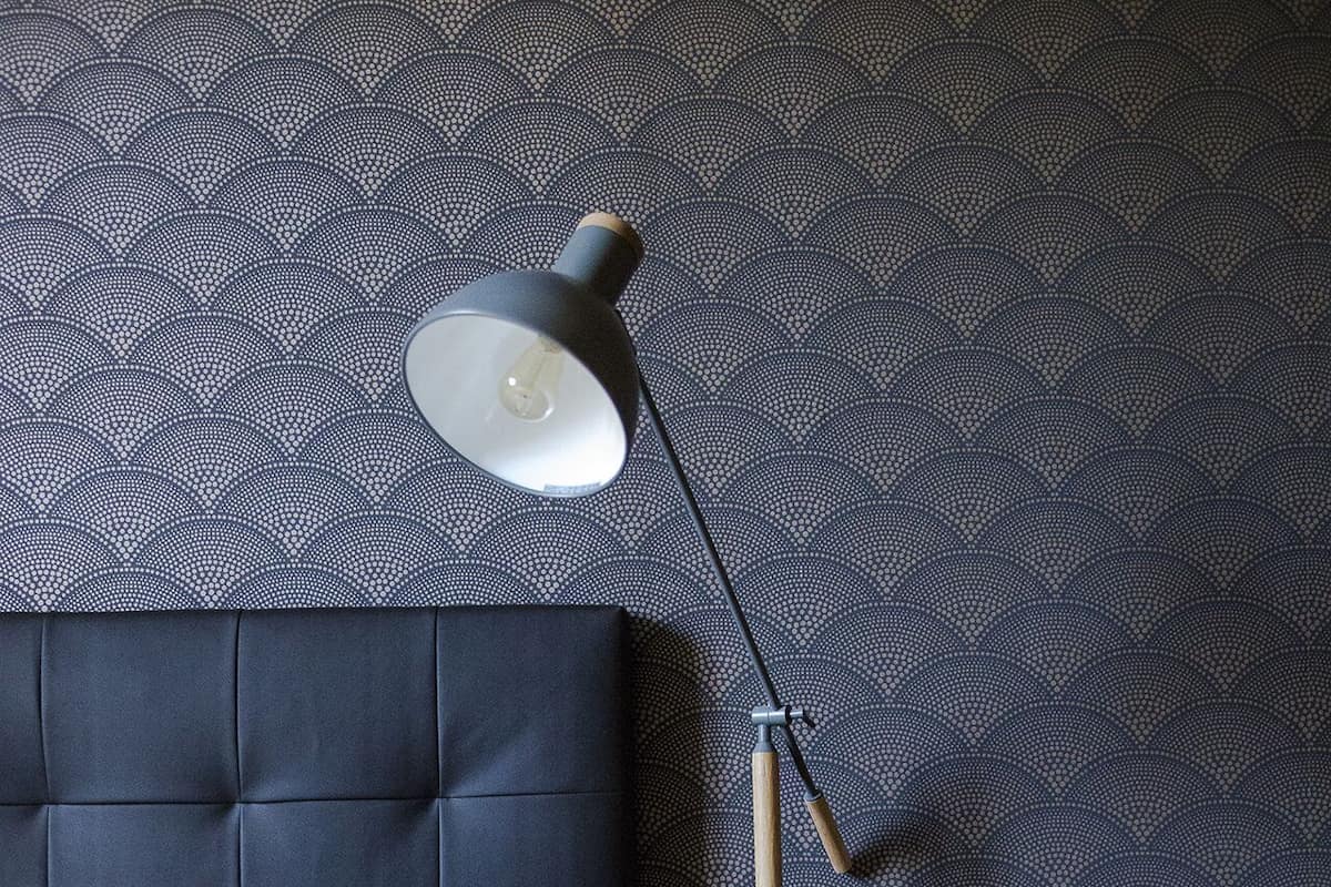 Una cama bien hecha con un cabecero oscuro, una lámpara de noche y un papel tapiz estampado.